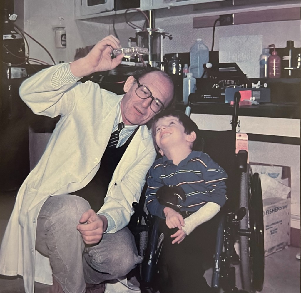 Dr. Baylink holding a culture plate up to the light beside a young boy in a wheelchair in his laboratory